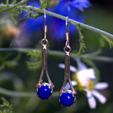 Pair of Lapis blue gemstone silver drop earrings hanging on a branch with blurred flowers in the background
